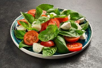 Fresh italian caprese salad with red tomato, green leaf basil, mozzarella cheese on a dark stone table.