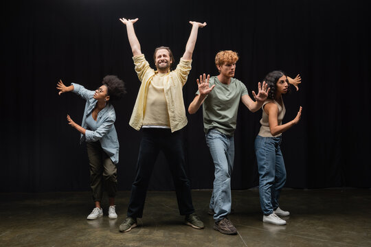 Young Man Standing With Raised Hands And Closed Eyes Near Multiethnic Students Rehearsing On Stage In Theater.