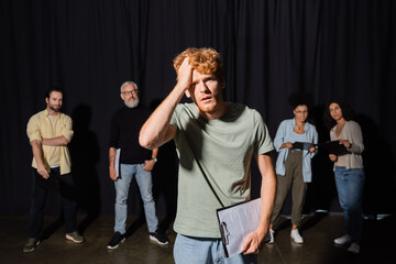frustrated redhead man holding clipboard and touching head while looking at camera near actors and...