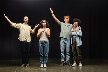 full length of happy multiethnic actors standing with raised hands on scene of theater.