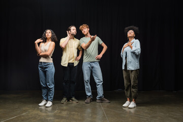 redhead man standing with hands on hips and pointing with hands near multicultural actors in acting studio.