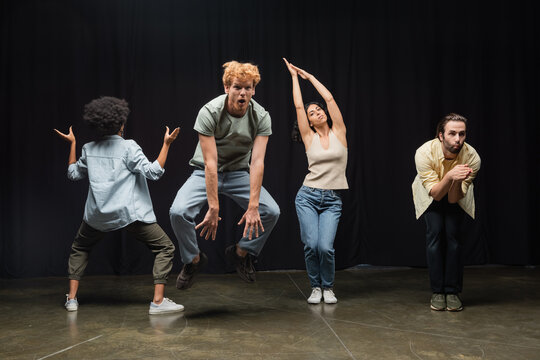 excited redhead man jumping near multiethnic actors posing during rehearsal in theater.