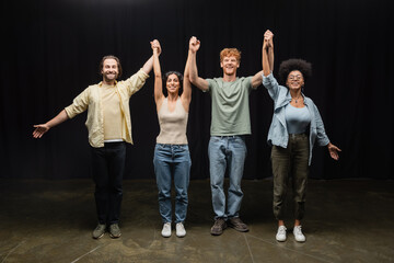full length of young and joyful interracial actors holding raised hands and smiling at camera in...