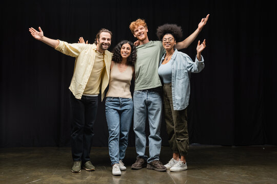 full length of cheerful interracial actors embracing while waving hands and smiling at camera in theater.