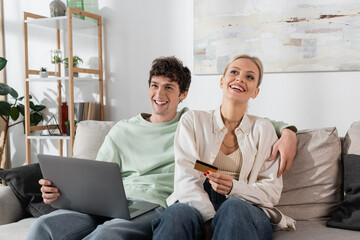 cheerful woman holding credit card near boyfriend with laptop doing online shopping.