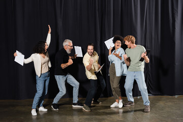 overjoyed multiracial theater troupe holding clipboards with screenplays and showing success gesture. Translation of tattoo: om, shanti, peace.