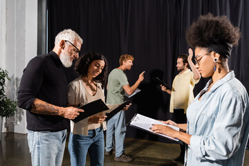 african american woman reading screenplay near bearded art director and troupe rehearsing in...