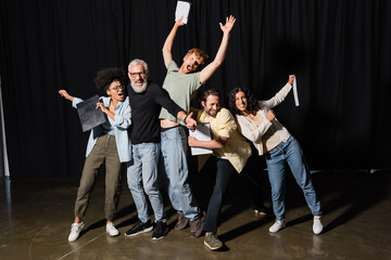 full length of excited interracial actors with bearded art director posing with clipboards on stage...