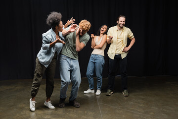cheerful interracial actors pointing at african american woman rehearsing with redhead man on stage in theater.