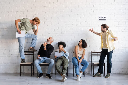 bearded man laughing and pointing at redhead man standing on chair near interracial actors waiting for casting.
