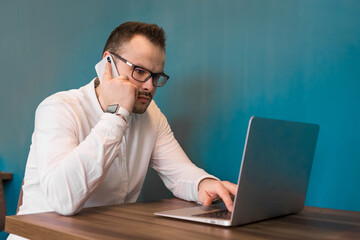 An adult guy in glasses and a white shirt, businessman sits at a table in a cafe in a laptop and solves business issues talking on a smartphone