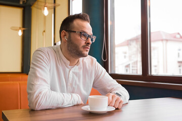 Young attractive businessman in a white shirt, glasses and headphones drinks coffee and looks thoughtfully out the window