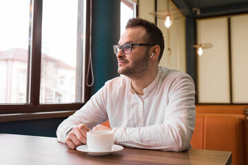 Fototapeta na wymiar Young attractive businessman in a white shirt, glasses and headphones drinks coffee and looks thoughtfully out the window