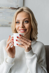 blonde young woman in white sweater holding cup of coffee and smiling at home.