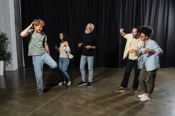 redhead man posing near excited multicultural actors and grey haired producer on stage in acting skills school.