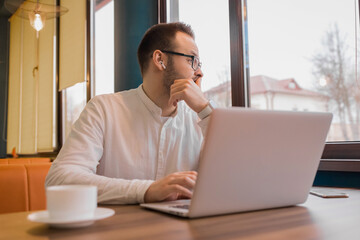 Young satisfied businessman sits in a laptop and looks out the window on a background of cafe interior