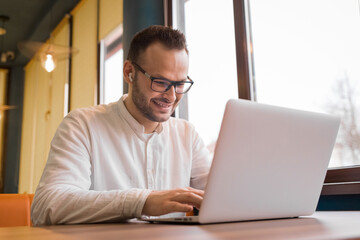 Funny, positive young guy businessman from Europe works in a laptop sitting at a table by the window