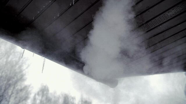 Slow motion video of steam from a chimney under the roof of a house in winter. Pipe from the sauna against the background of the sky and the roof of the building.