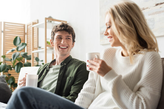 cheerful young man looking at blonde woman in sweater while holding cup with coffee in living room.