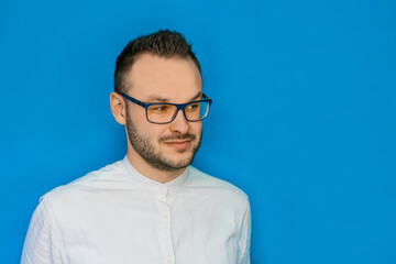 Obraz premium Portrait of a stylish young attractive guy with glasses and a white shirt looking to the side against a blue background
