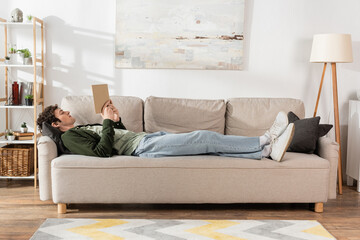 full length of young man with curly hair reading book while lying on couch in living room.