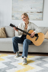 positive and blonde woman playing acoustic guitar while sitting on couch.