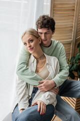 curly man hugging young blonde girlfriend in shirt and jeans.