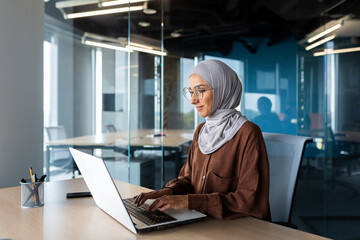 Successful smiling Arab woman in hijab working inside modern office, Muslim woman using laptop at work, business woman satisfied with achievement results typing on computer keyboard. - Powered by Adobe