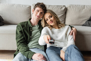 happy blonde woman clicking channels with remote controller near boyfriend in living room.
