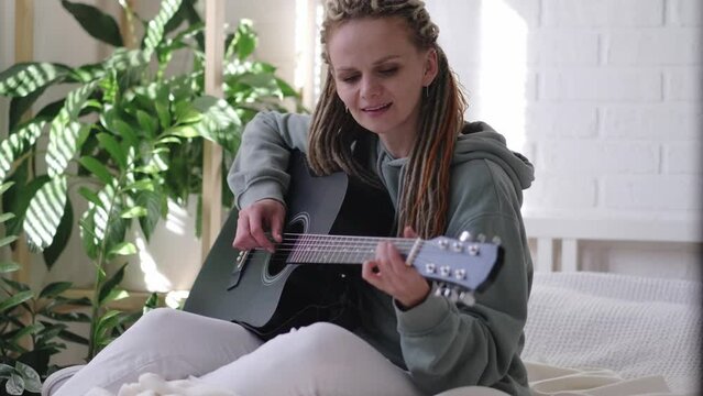 woman with dreadlocks on head singing song and playing acoustic guitar in sunny cottage bedroom, simple living and hobby relaxing
