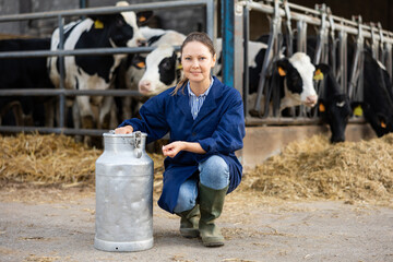 Cheerful woman farm worker squatting among rows of cow stables at milk can.