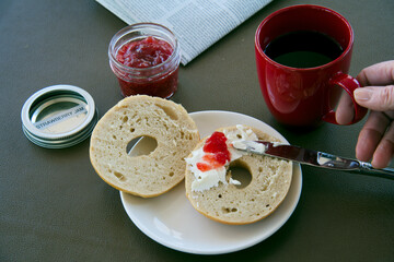 Lady's Hand spreading strawberry jam on creme cheese with newspaper and red coffee mug.
