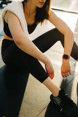 A young woman in black leggings and a top with a towel on her shoulder rests between exercises while sitting on the floor in the gym.