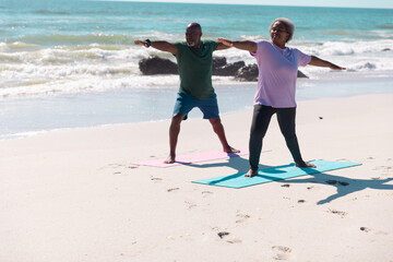 African american senior couple practicing warrior 2 pose on exercise mats at beach against sea