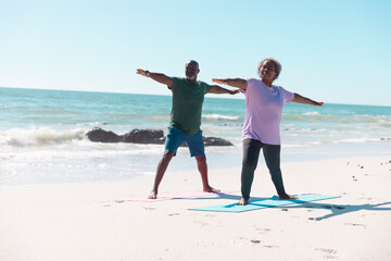 African american senior couple practicing warrior 2 pose on yoga mats at beach under clear sky