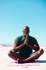 Bald african american senior man sitting cross legged and meditating at beach under clear blue sky