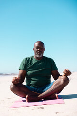 African american bald senior man with cross legged meditating on mat at beach against clear blue sky