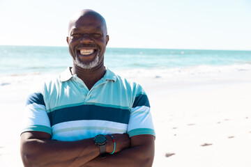 Smiling african american bald senior man with arms crossed standing against seascape and clear sky