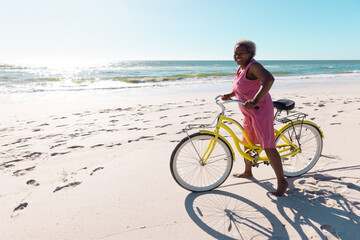 Fototapeta premium Carefree african american senior woman with bicycle standing on sandy beach against clear sky