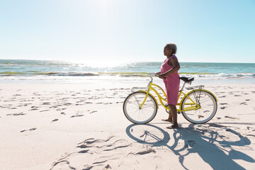 Side view of african american senior woman with bike standing on sandy beach against clear sky