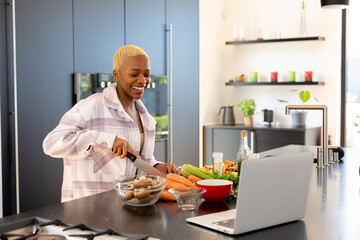 Happy african american woman cooking in kitchen, using laptop