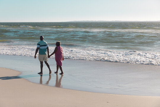 Rear view of african american senior couple holding hands and walking at seashore under clear sky