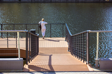 A woman stands and looks thoughtfully into a distance on a wooden pier on a jetty by a river, sea, lake in sunny day. Stainless steel railing, fence on a bridge. A girl walks alone in the street. - Powered by Adobe
