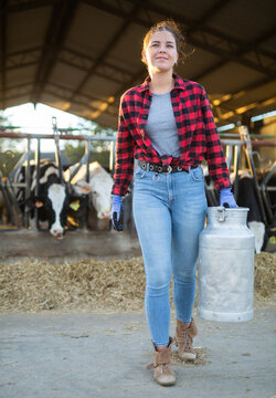 Full length portrait of cheerful young girl dairy farm worker carrying aluminum can of milk in cowshed against background of cows in stall