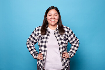 Young curvy woman smiling and posing with hands on hips, wearing a plaid shirt, isolated on a blue...