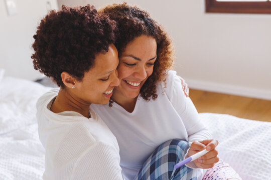 Happy diverse lesbian couple sitting on bed and looking at pregnancy test