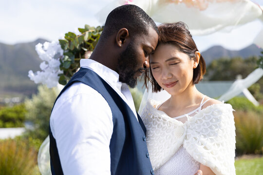 Happy, diverse bride and groom touching heads and smiling with eyes closed at outdoor wedding