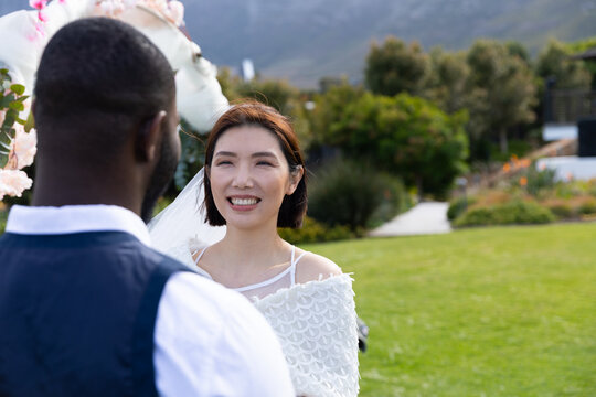 Happy asian bride smiling at diverse groom at outdoor wedding, copy space