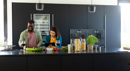 Happy diverse couple cooking, drinking wine and using tablet in modern kitchen, copy space