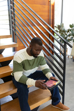 Vertical of african american man sitting on stairs at home using tablet computer, copy space